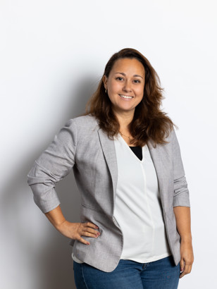 Natasha Pavic in een grijze blazer en witte blouse, staand met haar hand op de heup, tegen een neutrale achtergrond.