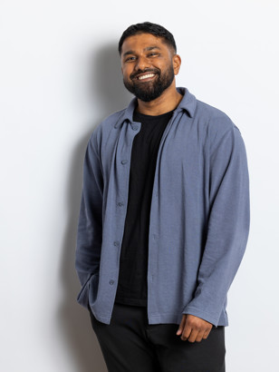 Wilkins Williams in een blauwe overhemdjacket met een zwart T-shirt, staand tegen een witte muur.