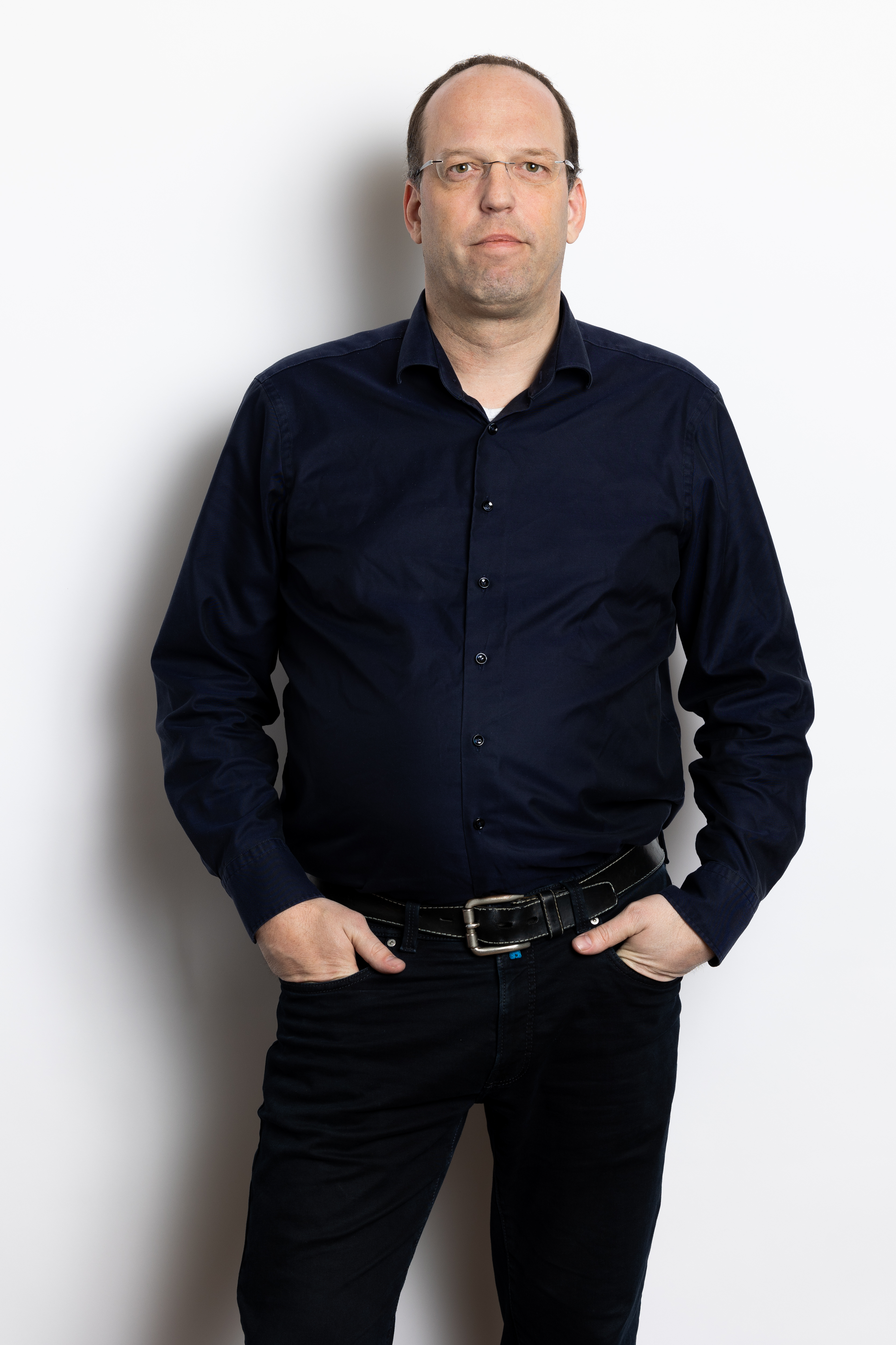 Paul Tromp in een donkerblauwe blouse met een zwarte riem, staand met één hand in zijn zak en de andere hand ontspannen naast zijn lichaam.