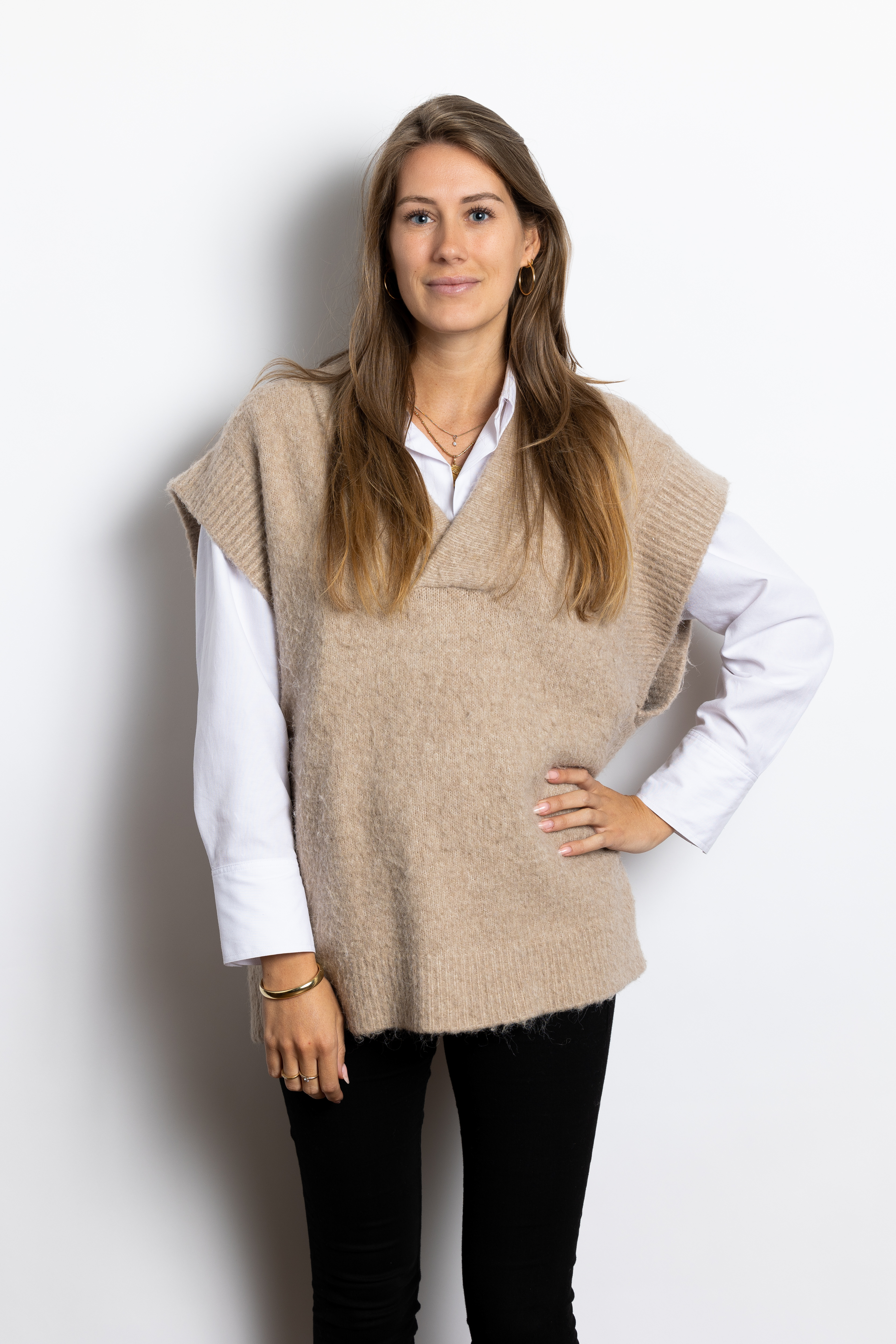 Ella Van Imhoff in een beige oversized trui met een witte blouse eronder, poseert met haar hand op haar heup tegen een witte achtergrond.