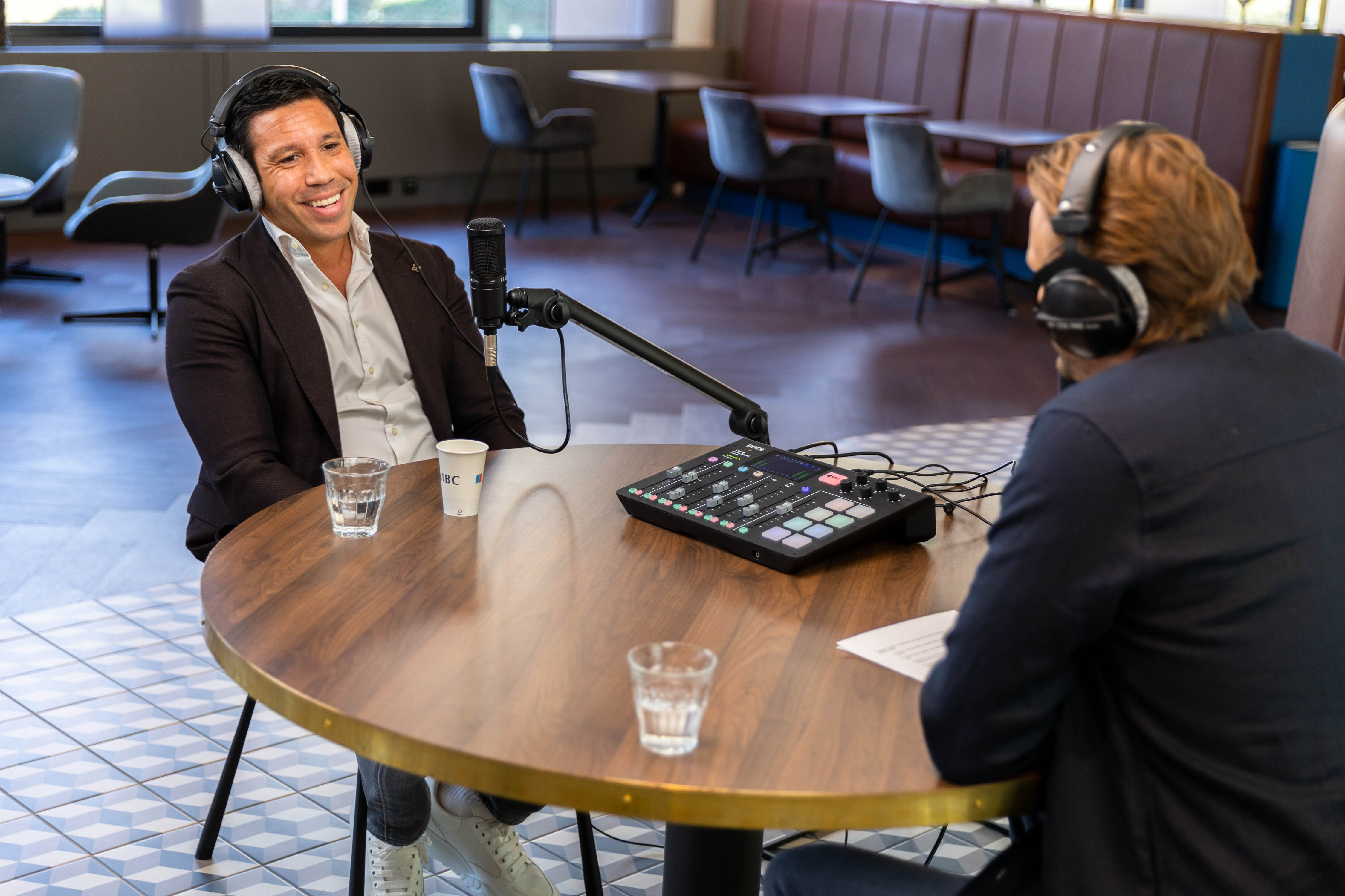 Twee mannen zitten aan een ronde tafel in een moderne ruimte; de ene spreekt in een microfoon terwijl de andere aandachtig luistert.