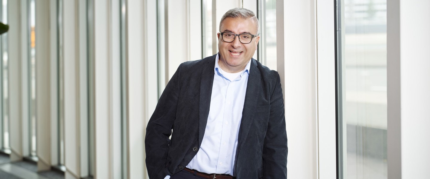 Een persoon in een zwart jasje en wit shirt leunt tegen een raam met verticale jaloezieën in een moderne kantooromgeving.