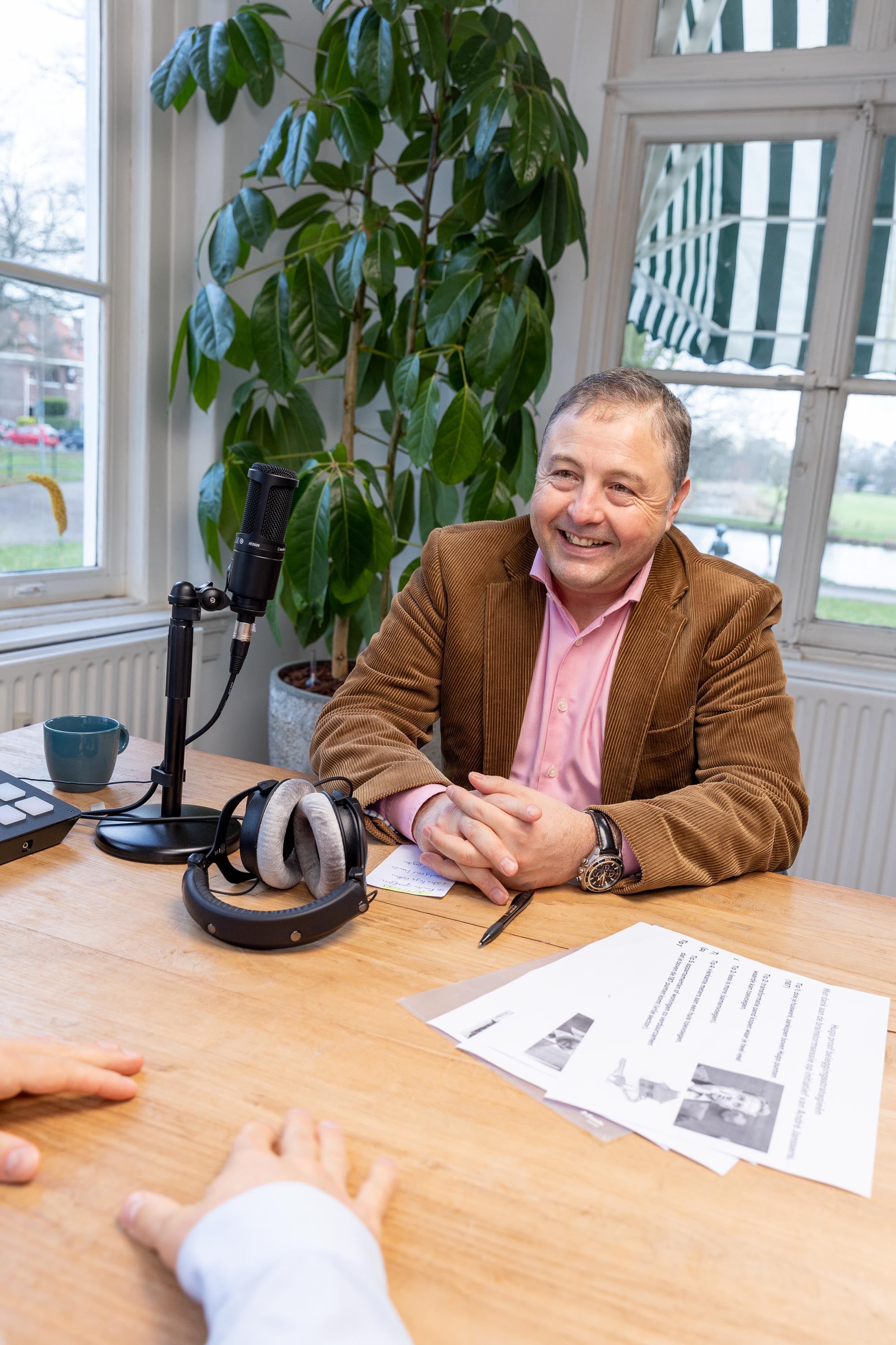 Een persoon in een bruine blazer zit aan een tafel met een microfoon en hoofdtelefoon, terwijl hij naar een andere persoon luistert.