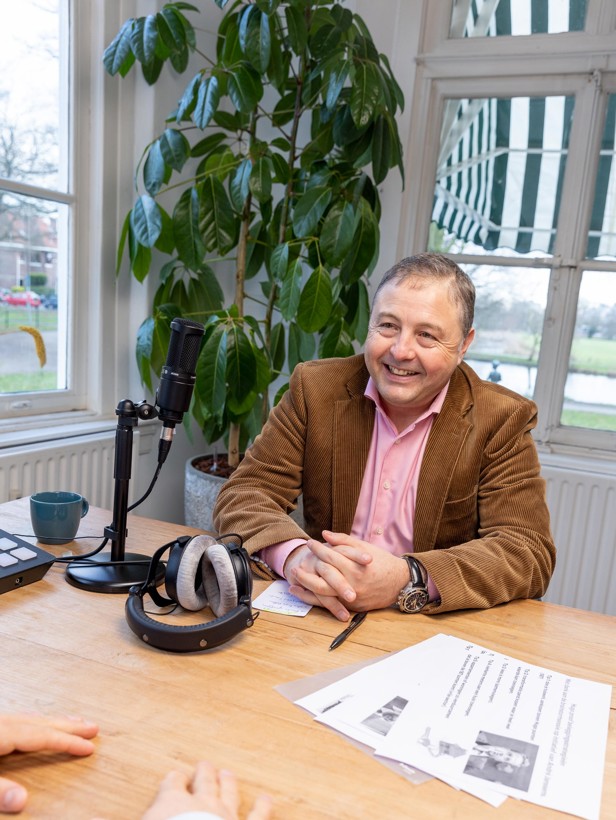 Een persoon in een bruine blazer zit aan een tafel met een microfoon en hoofdtelefoon, terwijl hij naar een andere persoon luistert.