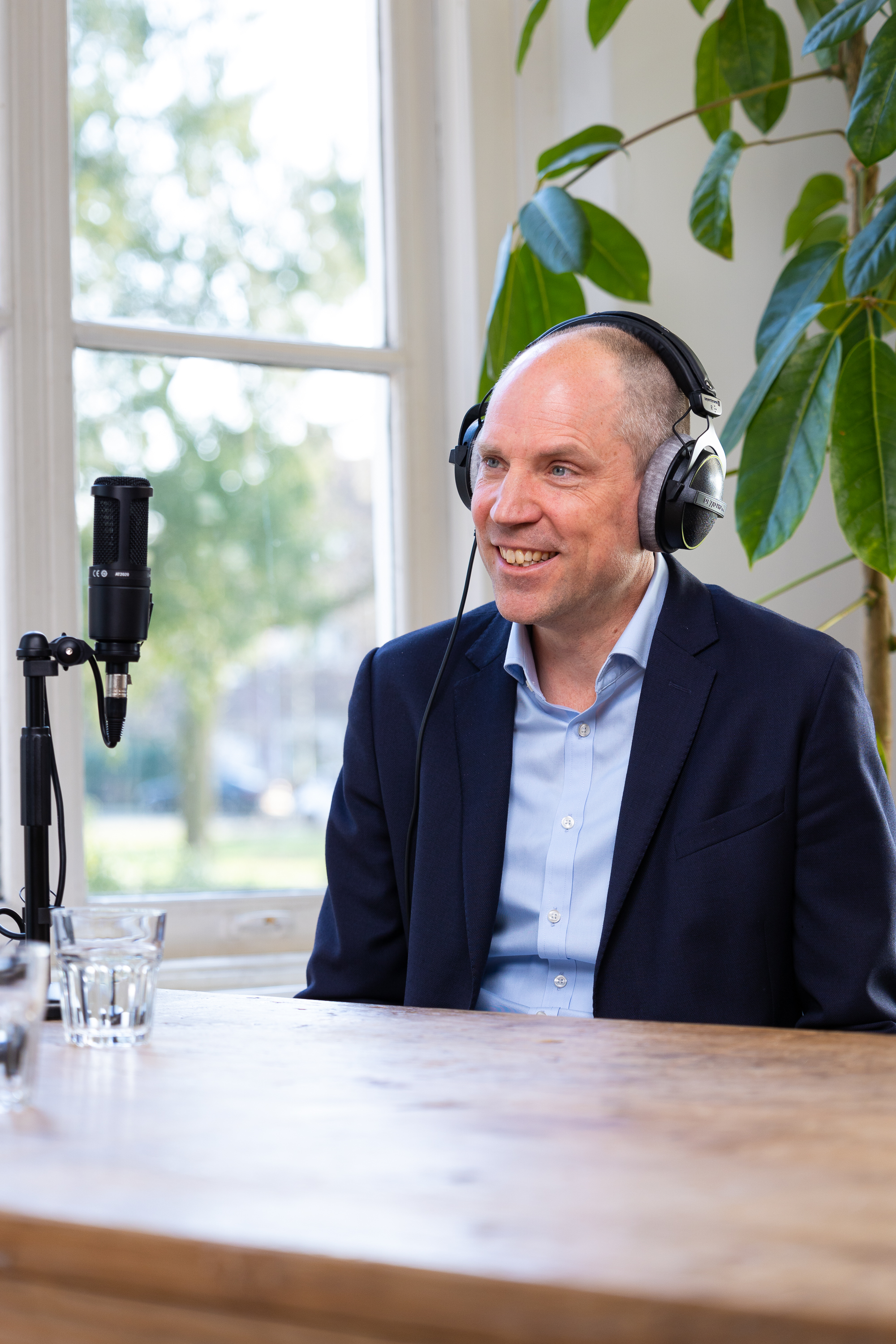 Dirk met een microfoon en hoofdtelefoon, zittend in een lichte ruimte met op de achtergrond enkele planten.
