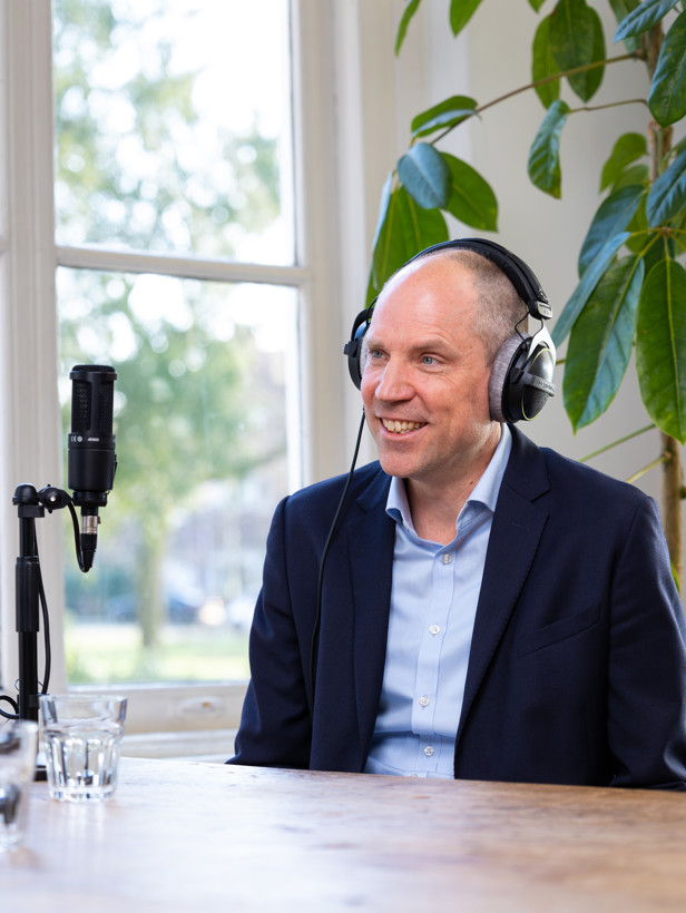 Dirk met een microfoon en hoofdtelefoon, zittend in een lichte ruimte met op de achtergrond enkele planten.