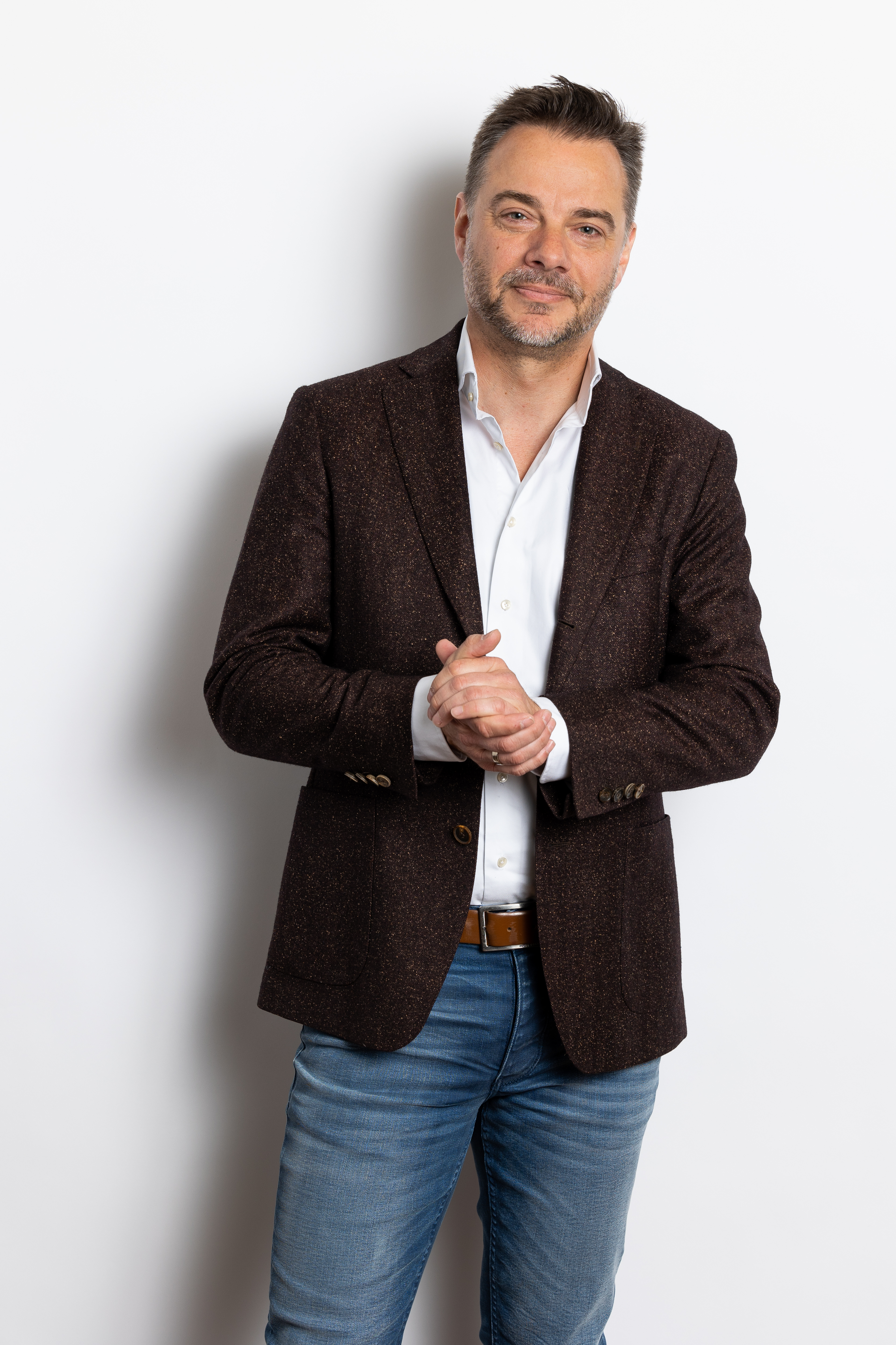 Sander in een donkerbruin colbert met gevouwen handen, staand voor een witte achtergrond.