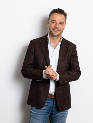 Sander in een donkerbruin colbert met gevouwen handen, staand voor een witte achtergrond.