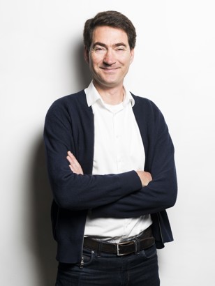 Frederik Berkhout met armen over elkaar, gekleed in een witte blouse en een donkerblauwe cardigan, staand tegen een witte muur.