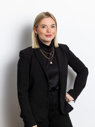 Carmen Oogjen in zwarte blazer en top, versierd met meerdere kettingen, poseert met een hand op haar heup tegen een witte achtergrond.