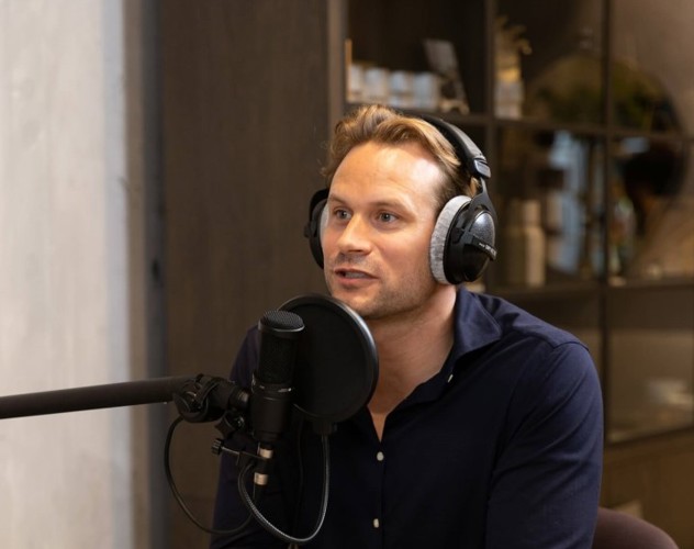 Dirk Taat in een zwart overhemd met een hoofdtelefoon zit voor een microfoon in een moderne studio, terwijl hij geïnterviewd wordt.