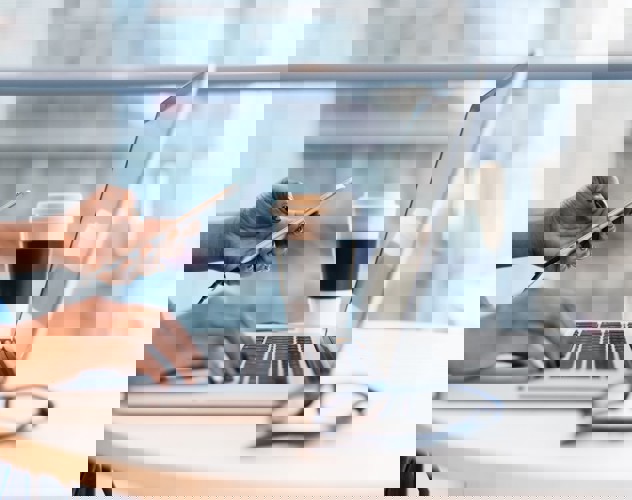 Een persoon met zwart gelakte nagels typt op een laptop terwijl hij/zij een smartphone vasthoudt, met een koffiebeker naast zich.