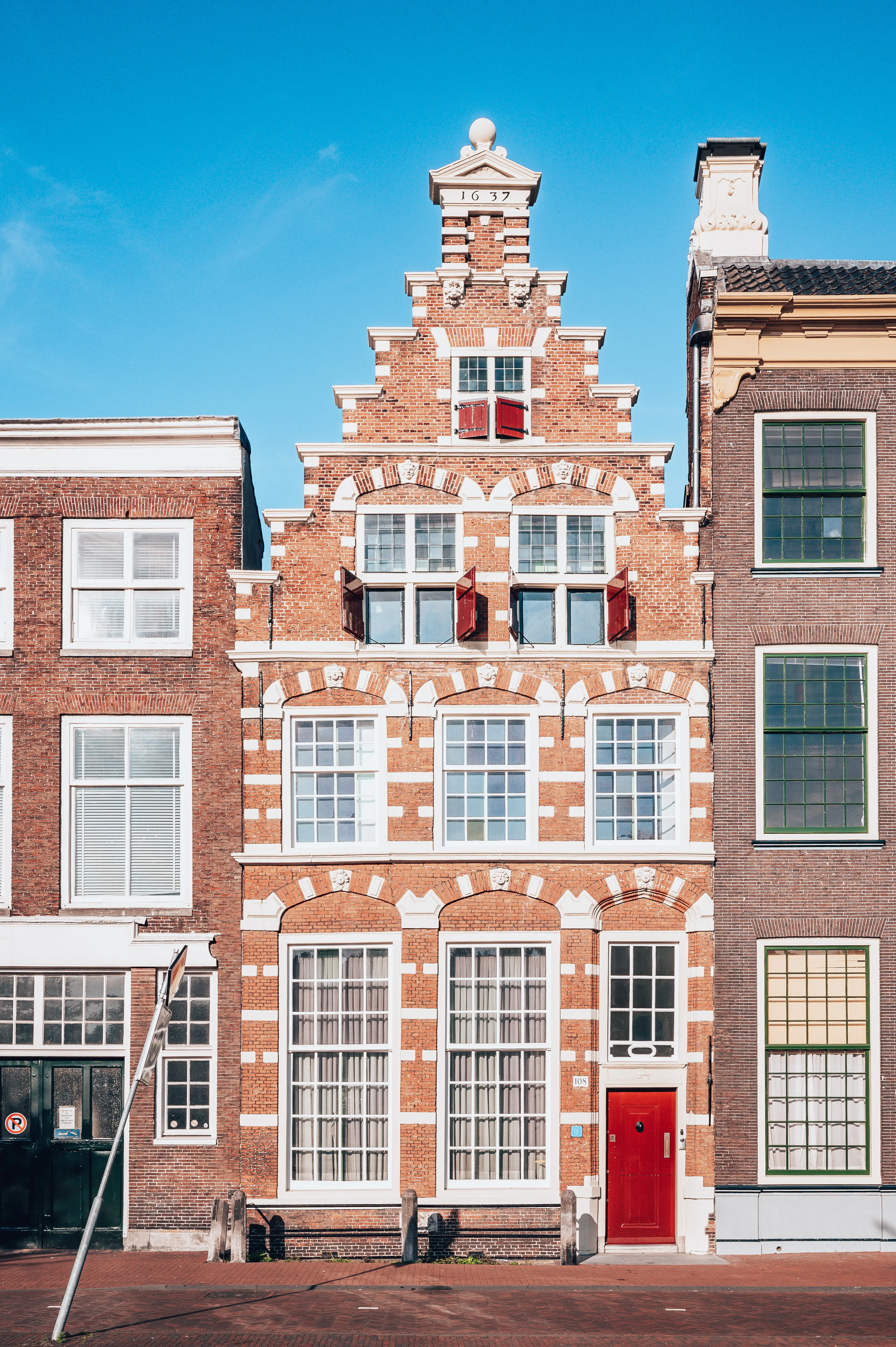 Roodstenen gebouw met witte accenten en een gedetailleerde gevel, omringd door twee moderne gebouwen.