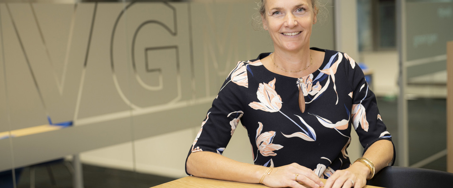 Vrouw in een bloemenblouse zit aan een tafel in een modern kantoor.