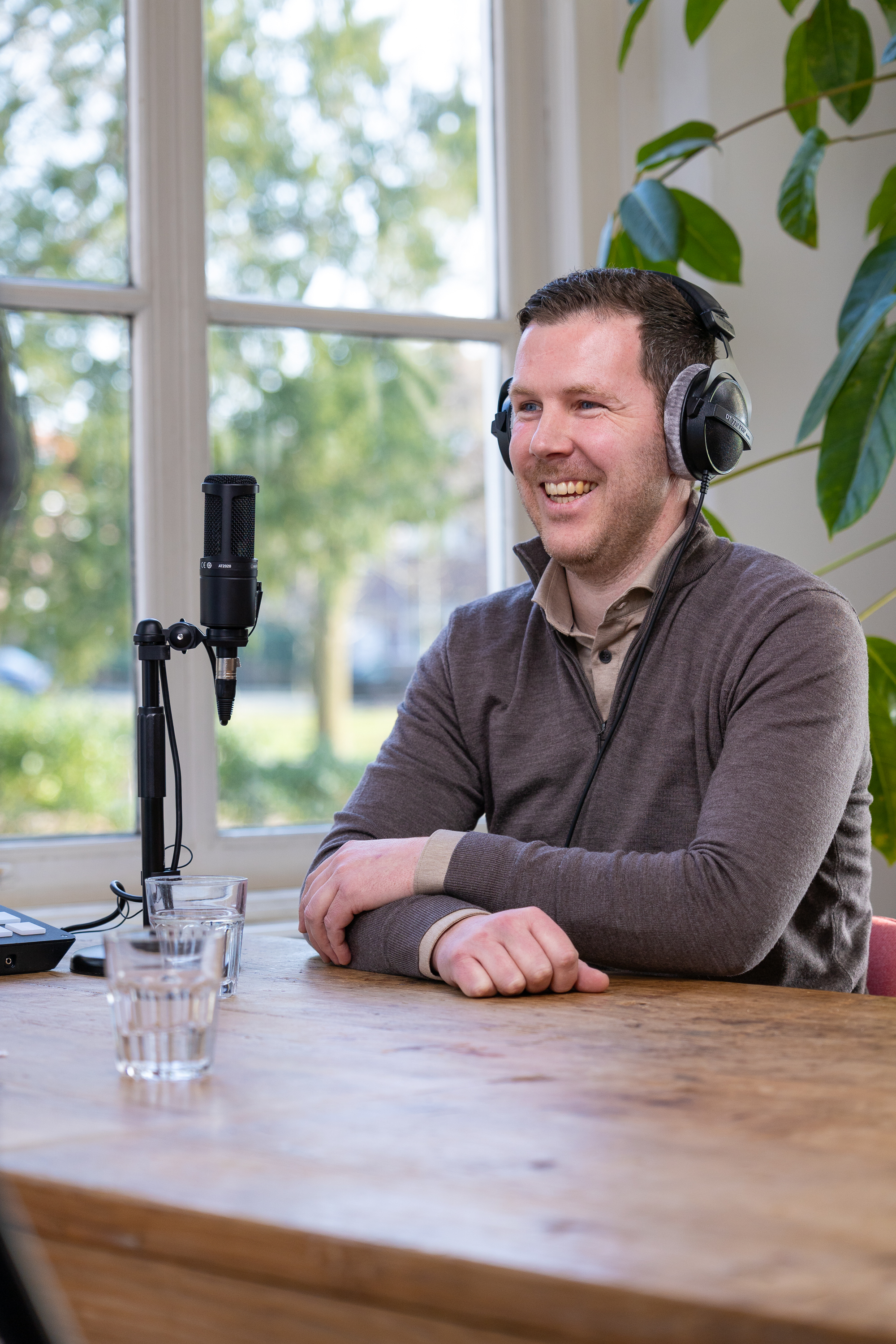 Sven met een microfoon en koptelefoon, leunend tegen een raam met groene planten op de achtergrond.
