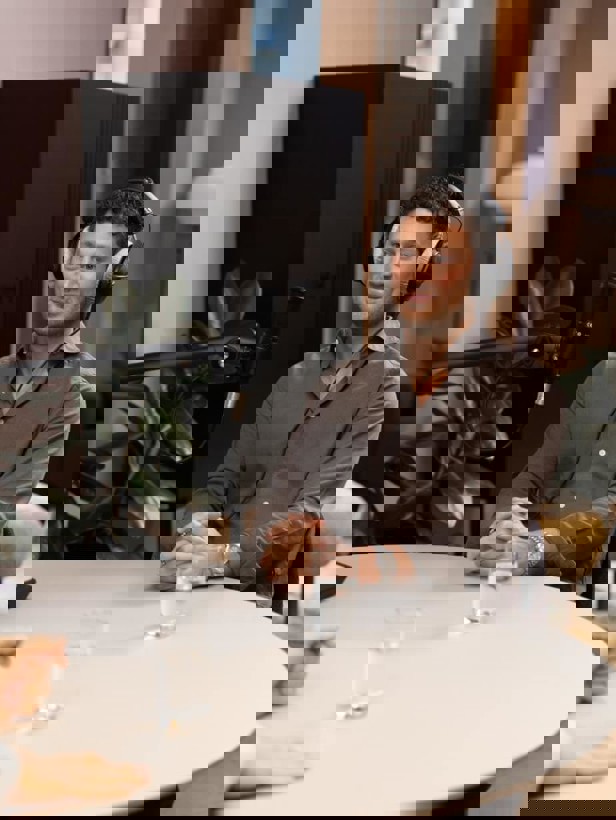 Een persoon met een hoofdtelefoon zit aan een tafel met een microfoon naar hem gericht. Op de tafel staan twee glazen water.