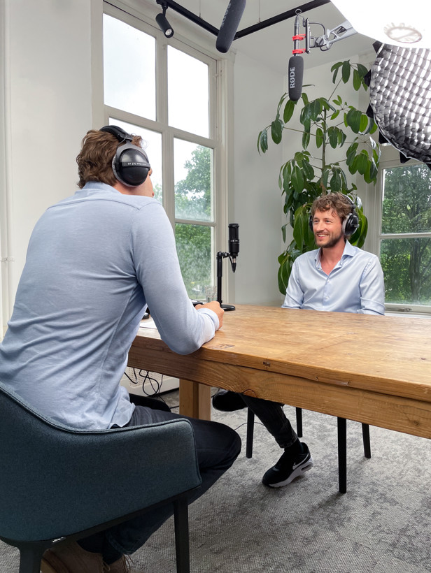 Twee personen zitten aan een houten tafel in een lichte ruimte, terwijl opnameapparatuur is opgesteld voor een gesprek.