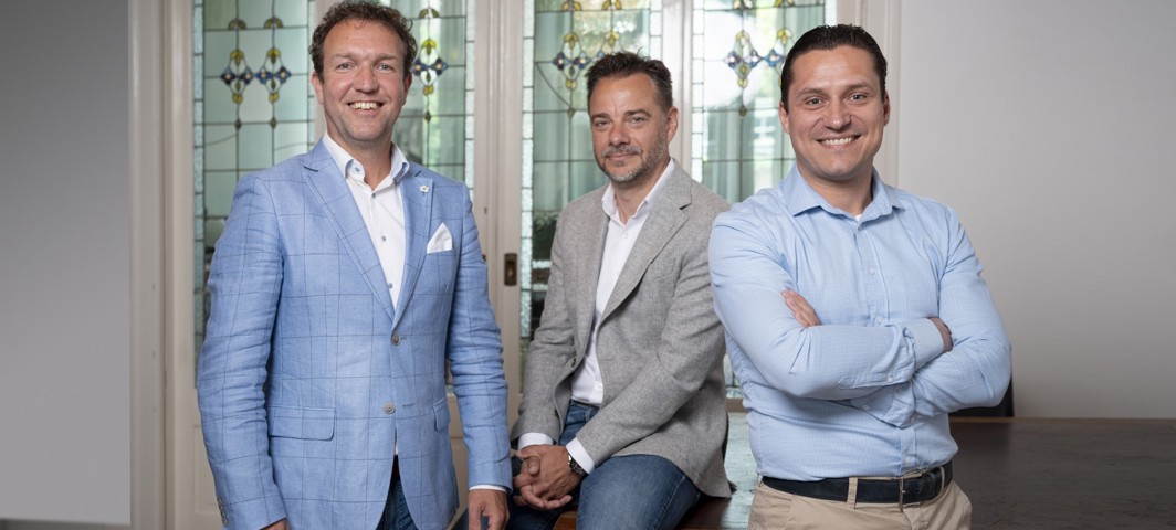 Drie mannen in zakelijke kleding staan en zitten voor een decoratieve glazen wand met loodramen.