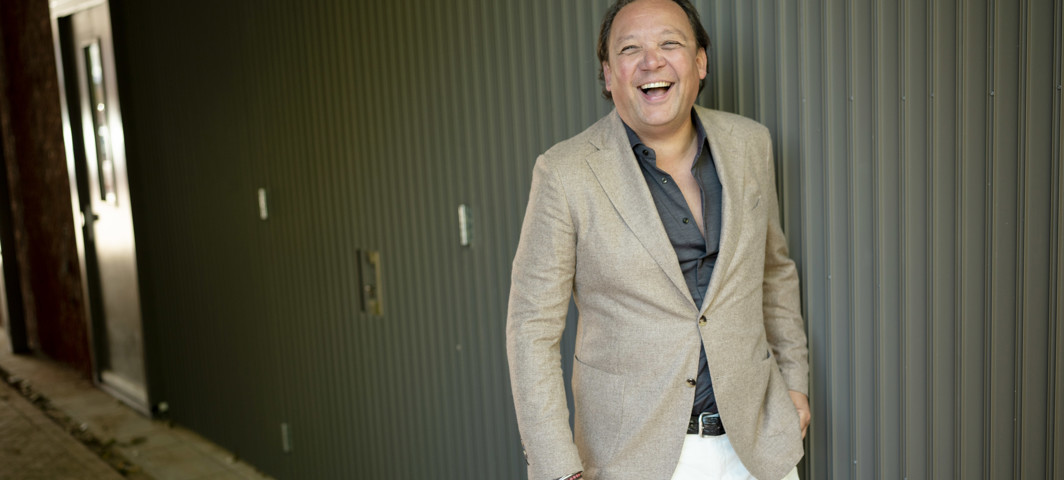 Man in een lichtgrijze blazer en zwart shirt poseert tegen een beklede muur.