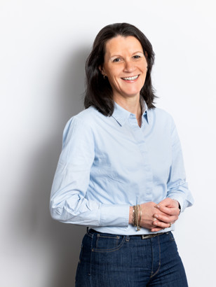 Marina Buttingha Wichers in een lichtblauwe blouse en donkere jeans, met haar handen voor haar gevouwen, staat voor een witte achtergrond.