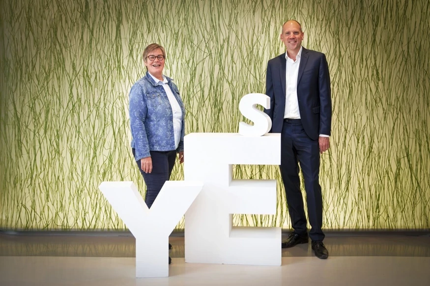 Twee mensen staan naast grote witte letters die samen 'JA' vormen, tegen een groene, grafische achtergrond.