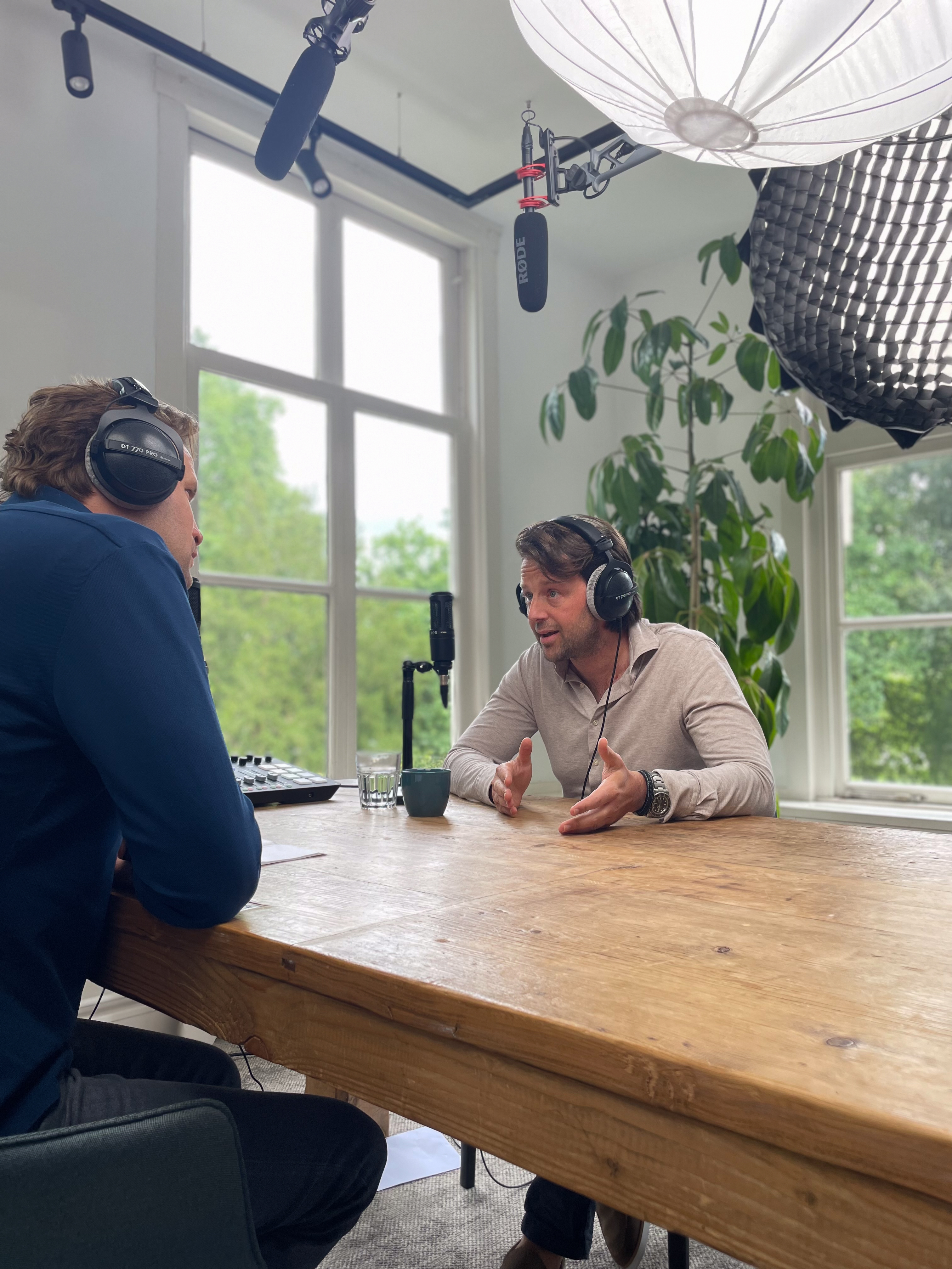 Twee mannen converseren tijdens een podcastopname, met een microfoon en een groene plant op de achtergrond.