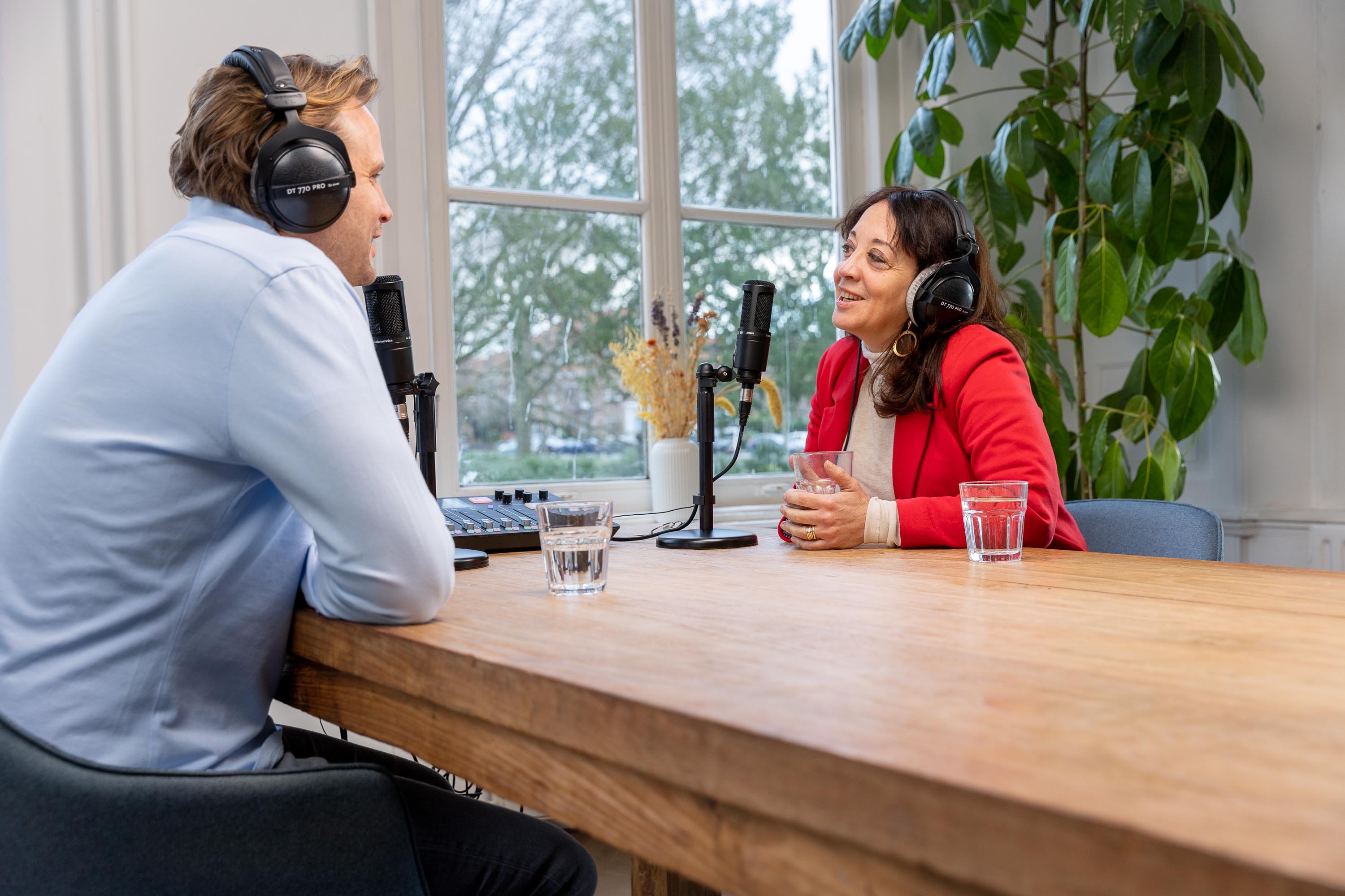 Twee mensen converseren aan een houten tafel, beiden met een microfoon en hoofdtelefoons, in een lichte ruimte omringd door planten.