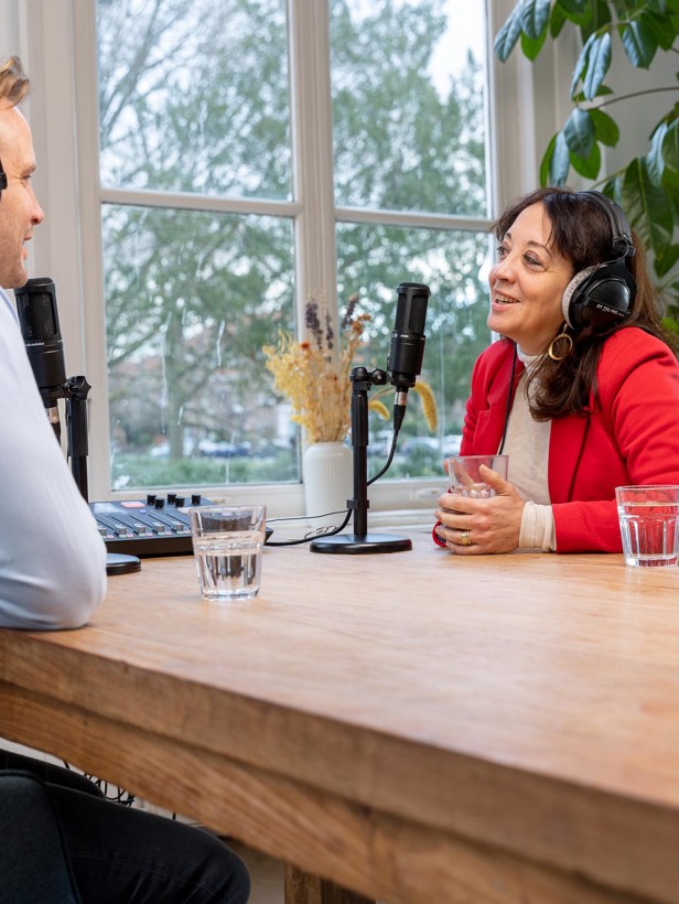 Twee mensen converseren aan een houten tafel, beiden met een microfoon en hoofdtelefoons, in een lichte ruimte omringd door planten.