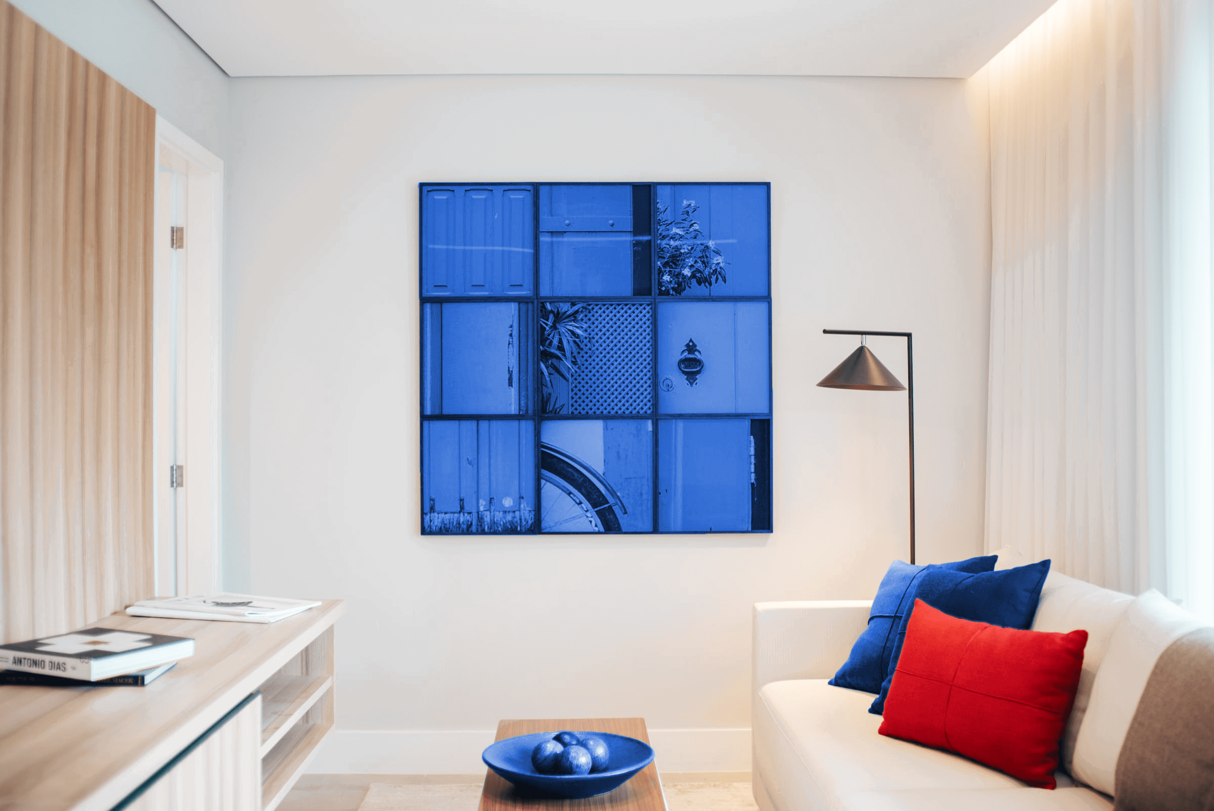 Een moderne woonkamer met een blauw kunstwerk aan de muur, een witte bank met blauwe en rode kussens, en een lamp in de hoek.