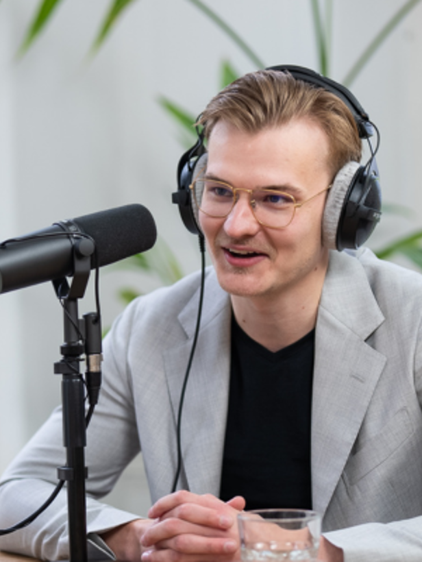 Jelle in grijze blazer en koptelefoon, zittend aan tafel met microfoon, geconcentreerd op een gesprek. Groene plant op de achtergrond.