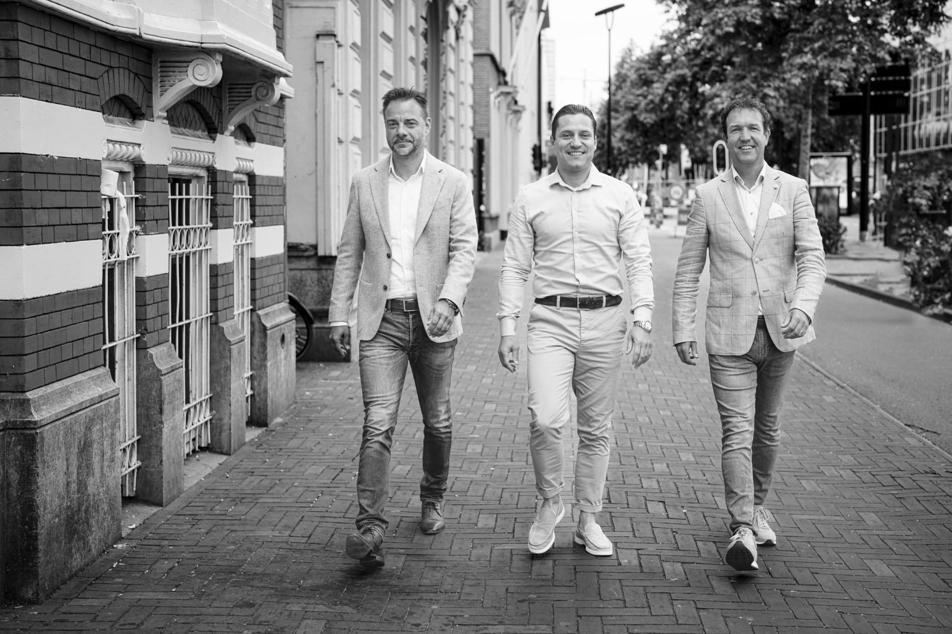 Drie mensen in formele kleding wandelen over een trottoir langs historische gebouwen in zwart-wit.