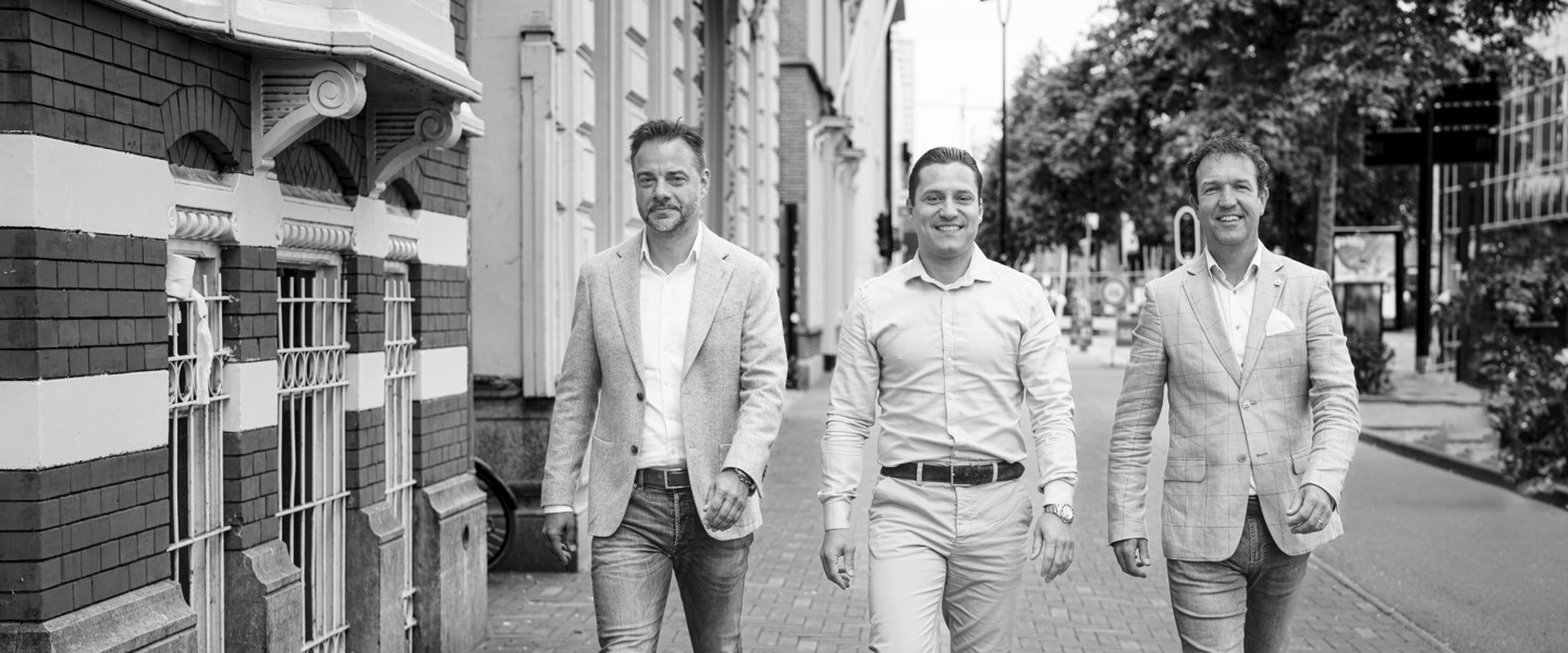 Drie mensen in formele kleding wandelen over een trottoir langs historische gebouwen in zwart-wit.