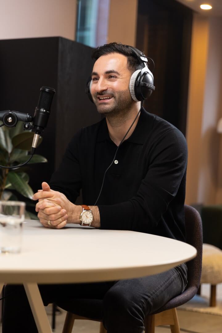 Onur in zwart T-shirt en met hoofdtelefoon, zittend bij een microfoon in een moderne setting.