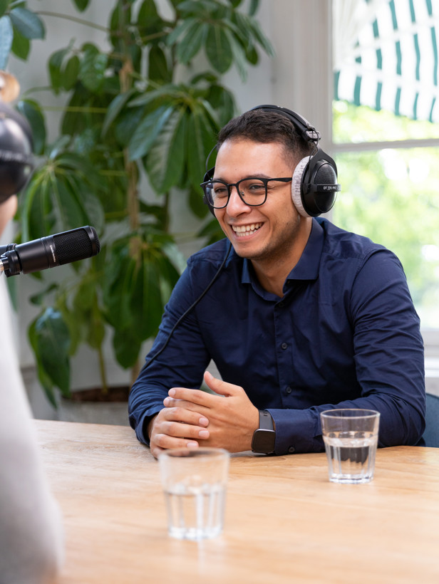 Twee mensen zitten aan een houten tafel, met een microfoon, hoorapparatuur en glazen water, terwijl ze een gesprek of interview voeren.