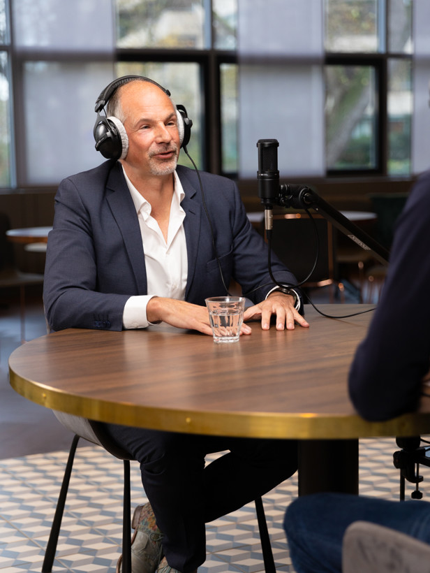 Man in pak met een headset, zittend aan een tafel met een glas water, in gesprek met iemand buiten beeld.